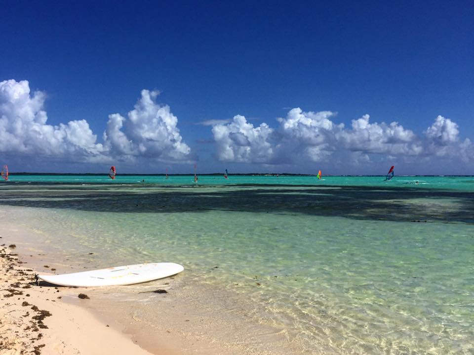A white board on a sandy beach beside clear turquoise water, with windsurfers beneath a blue sky.
