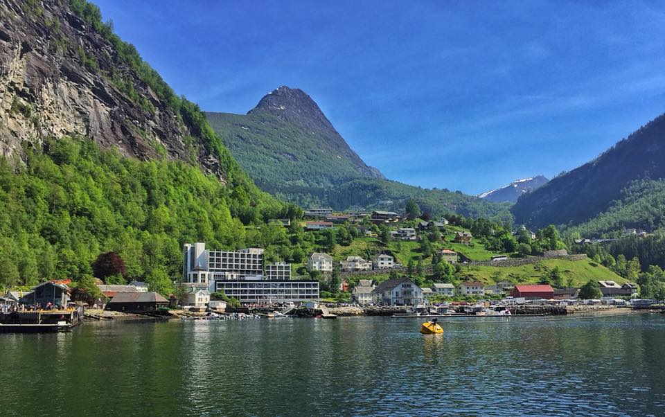 A waterfront village beneath green mountains, viewed across a calm fjord.