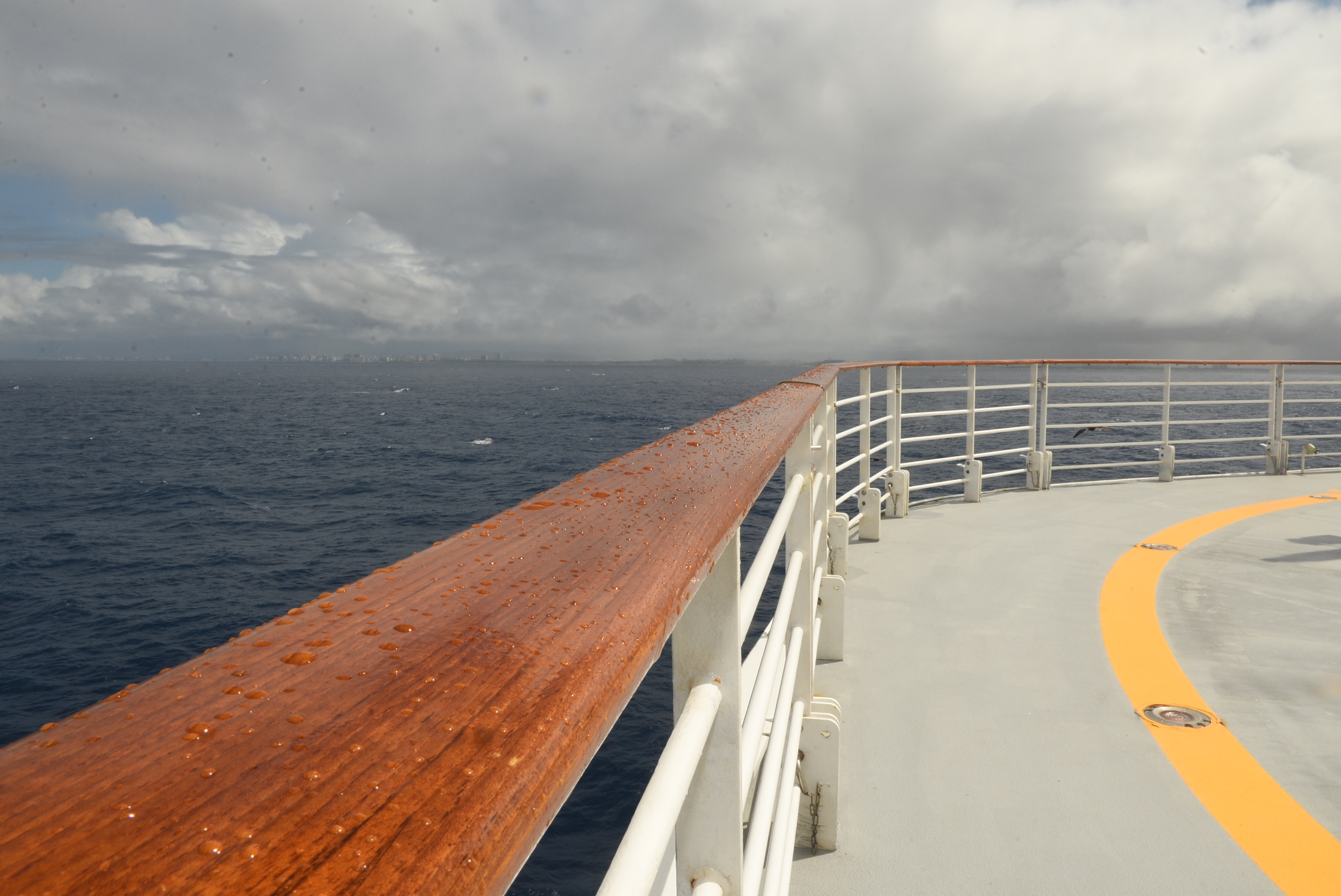 A wooden cruise-ship railing overlooking the ocean beneath a cloudy sky.